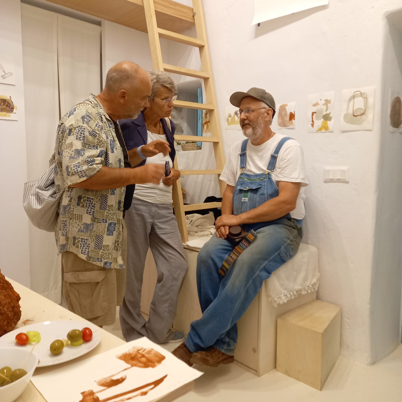Jeff Benjamin, in a cap and overalls, sitting and talking with a visitor in the studio