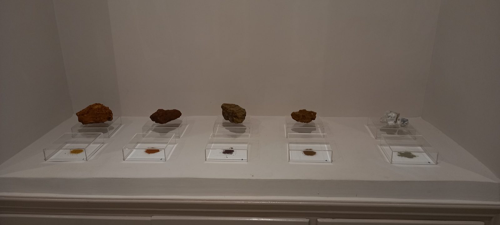 A row of framed earth-pigment drawings by Jeffrey Benjamin on a white wall of the Archaeological Museum of Serifos, beside the wall text