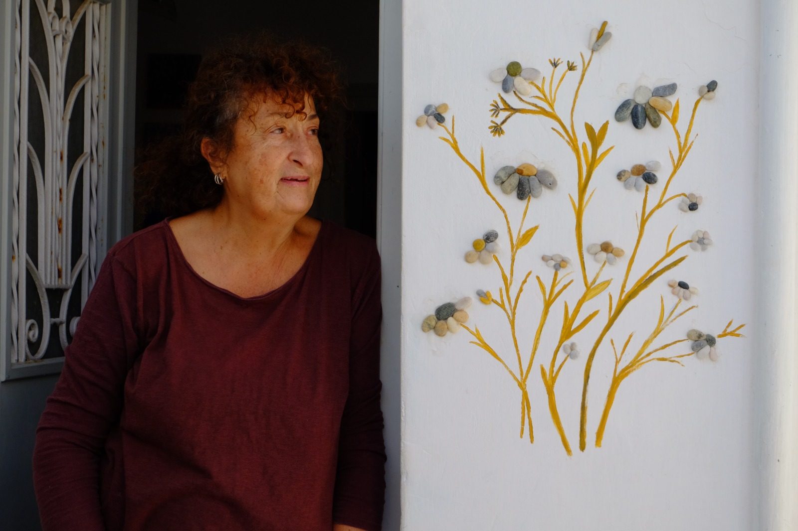 Kiki Zachariou at a doorway beside a wall painted with flowers made of sea pebbles